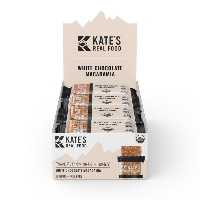 A display box of Kates Real Food Organic Energy Bar - White Chocolate Macadamia includes twelve gluten-free bars with organic oats and whole-food ingredients, each bar individually wrapped and labeled as USDA organic.