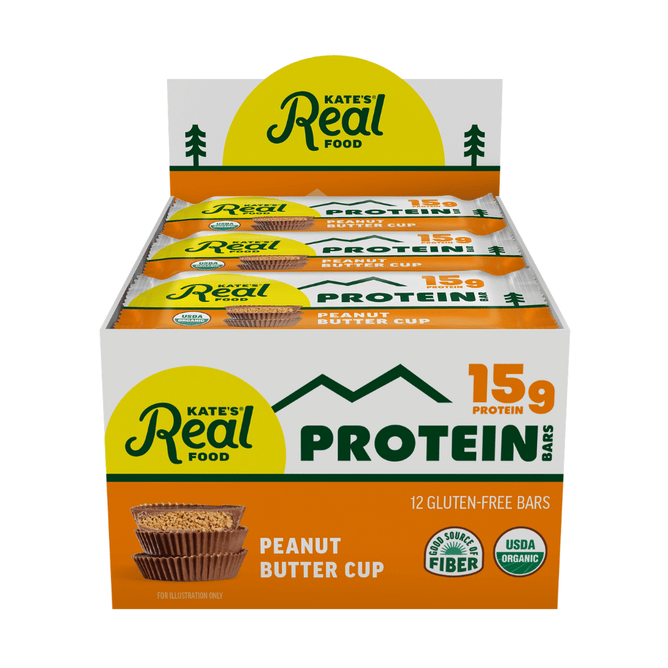 A box of Kate’s Real Food Organic Protein Bars, labeled 15g protein and containing 12 gluten-free bars, showcases organic and soluble fiber certifications on the front. Made with whole-food ingredients for clean fuel, the packaging displays illustrations of two bars and peanut butter cups.