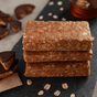 Three Kate’s Real Food Organic Protein Bars stacked on a dark slate surface, with scattered oats and chocolate-covered treats in the background—a delicious display of clean fuel crafted from wholesome, whole-food ingredients.