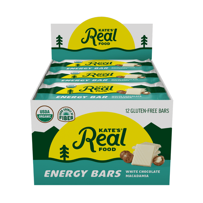 A display box of Kate’s Real Food Organic Energy Bars in White Chocolate Macadamia flavor, containing 12 gluten-free bars made with whole-food ingredients such as oats and honey. The box features a yellow mountain graphic and carries the USDA Organic label, as well as a good source of fiber designation.