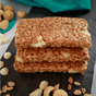 Three Kate’s Real Food Organic Energy Bars with visible macadamia nuts are stacked on a dark slate surface, surrounded by scattered nut pieces and a green cloth in the background—these bars blend oats and honey to deliver clean, on-the-go energy.