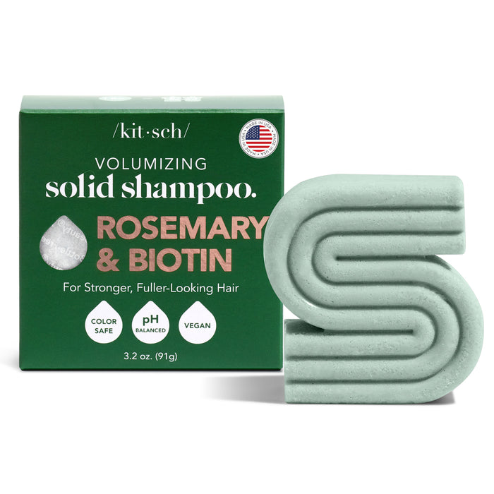 A green box labeled Volumizing Solid Shampoo: Rosemary & Biotin stands next to a matching green, S-shaped solid shampoo bar. The box highlights features like color safe, pH balanced, and vegan.