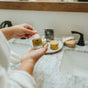 A person holds a small bottle labeled “Nectar Serum with Natural Retinol and Tallow” from Hearth and Homestead over a marble bathroom counter, with skincare products, a brush, sandals, and a black faucet visible in the background.
