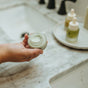 A person holds an open jar of Hearth and Homestead Tallow Face Cream - Anti-Aging over a marble bathroom sink, with two dropper bottles on a tray in the background.
