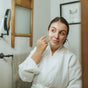 A woman in a white bathrobe applies Hearth and Homestead’s Tallow Face Cream - Anti-Aging to her skin while looking into a mirror in a bathroom.