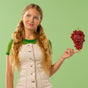 A young woman with long blonde hair, in a beige dress over a green shirt, smiles while balancing red grapes—evoking MegaFood Kids One Daily Multivitamin Soft Chews—on her finger against a light green background.