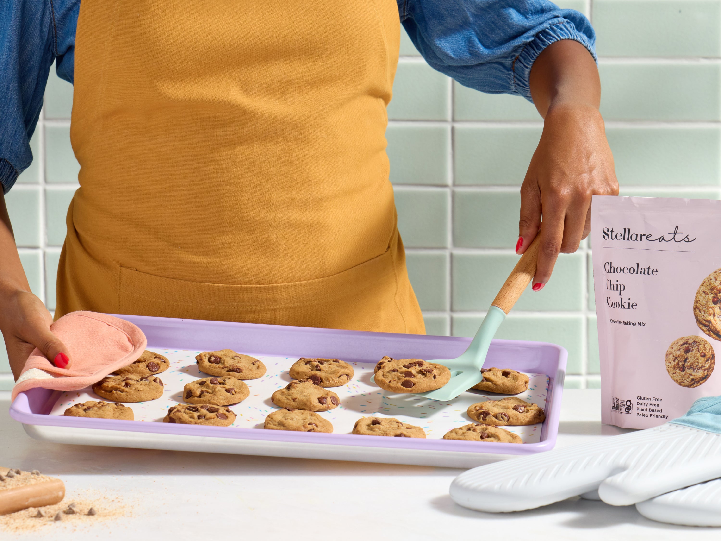 A person wearing a yellow apron uses a spatula to lift chocolate chip cookies from a baking tray. A bag labeled Stellareats Chocolate Chip Cookie is on the counter beside oven mitts.