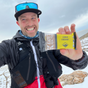 A smiling man in outdoor gear holds up a Kates Real Food Organic Energy Bar - Lemon Coconut, an energizing snack, as he stands in a snowy mountain landscape.