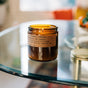 A Large Concentrated Soy Candle from P.F. Candle Co. rests on a glass table, its amber jar and nearby lid capturing the warm glow of the soy wax flame, set against a gently blurred background.