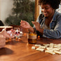 Two people sit at a wooden table playing a game with dice and dominoes. A Large Concentrated Soy Candle by P.F. Candle Co. in an amber jar and a glass of red wine rest on the table. One person smiles and gestures with their hands, adding warmth to the cozy scene.
