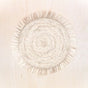 The LIKHA Raffia Fringe Placemats, set of 2, are round beige placemats made from natural raffia and handwoven by Filipino artisans, featuring a stylish fringe border. Displayed on a light wooden surface.