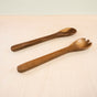 Two Fair Trade utensils from LIKHAs Acacia Essential Serving Set rest on a light-colored surface: a smooth wooden spoon and a three-pronged spork.
