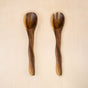 The LIKHA Acacia Essential Serving Set with Wiggly Handles features two ethically made wooden utensils—one spoon and one forked spoon—perfect for serving salads on any light wooden surface.