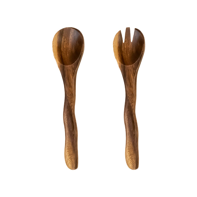 On a white background: LIKHA’s Acacia Essential Serving Set with Wiggly Handles, handcrafted from sustainably sourced acacia wood—a spoon on the left and a matching two-pronged fork on the right.