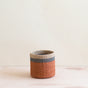 The LIKHA Abaca Cylinder Tabletop Basket, ethically crafted from Manila hemp fiber with orange, gray, and beige bands, rests on a light wooden surface against a simple pale background.