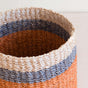 A close-up of the LIKHA Abaca Cylinder Tabletop Basket, ethically crafted from Manila hemp fiber with beige, blue, and coral bands, showcasing its textured surface and open top against a neutral background.
