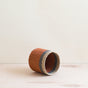 The LIKHA Abaca Cylinder Tabletop Basket, expertly woven from Abaca fiber in orange, beige, and gray stripes, rests on its side on a light wooden surface against an off-white background.
