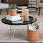A round black coffee table with books, notebooks, and a small potted plant sits beside the LIKHA Abaca Cylinder Tabletop Basket filled with greenery and a magazine, all arranged on a patterned rug in a cozy living room.