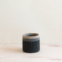 The LIKHA Abaca Cylinder Tabletop Basket is a small, handcrafted cylindrical basket with dark gray sides and a light beige rim, made from Abaca fiber. Its shown on a light wooden surface against a neutral background.