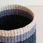 A close-up of the LIKHA Abaca Cylinder Tabletop Basket, handcrafted from abaca fiber with beige, gray, and dark blue horizontal stripes, displayed on a light-colored surface.