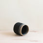 The LIKHA Abaca Cylinder Tabletop Basket, handcrafted from black and natural tan Abaca fiber, rests on its side atop a light wooden surface with a plain, off-white background.