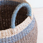 A close-up of the LIKHA Abaca Bulge Basket shows brown, beige, and blue stripes with a rounded blue handle on the rim. The ethically made woven abaca fibers display exquisite texture and detail.