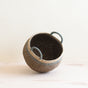 The Abaca Bulge Basket by LIKHA is a round, ethically handwoven brown basket made from Manila hemp, featuring gray or blue-tinted handles and rim. It sits tilted on a light wooden surface against a plain off-white background.
