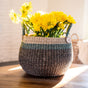 The LIKHA Abaca Bulge Basket, handwoven from ethically sourced Manila hemp with green trim, holds bright yellow flowers and rests on a sunlit wooden surface.