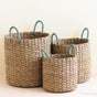 The LIKHA Seagrass Cylinder Basket with Handles, a nested set of 3 ethically made baskets, features two sky blue handles each and is displayed on a light surface against a pale background.
