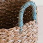 A close-up shows the LIKHA Seagrass Cylinder Basket with Handles, a nested set of 3, featuring textured handles wrapped in sky blue and natural fibers. The ethically made basket sits elegantly on a light surface.