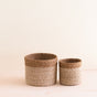 Two LIKHA Abaca Cylinder Tabletop Baskets, a nested set of 2 in natural brown, are displayed side by side on a light wooden surface with a plain beige background.