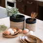 Two LIKHA Abaca Cylinder Tabletop Baskets, nested set of 2, sit on a kitchen counter—one holds wooden utensils, the other a leafy plant. Wooden plates with brown and white eggs are arranged in front.