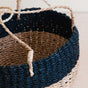 A close-up of the LIKHA Abaca Round Hanging Planter, handwoven from natural abaca fiber with beige and dark blue stripes and thick, twisted rope handles, shown on a light wooden surface.