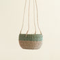 The LIKHA Abaca Round Hanging Planter, handwoven in a green and beige pattern from abaca fiber, hangs by three thin rope handles against a plain light background.