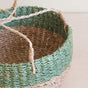 A close-up of the LIKHA Abaca Round Hanging Planter, handwoven from natural and green Abaca fibers with two braided handles, displayed on a light wooden surface.