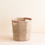 The LIKHA Abaca Octagon Basket, crafted ethically from natural straw, has a geometric cylindrical shape with two handles wrapped in light pink thread. Its displayed on a light wooden surface against a neutral background.