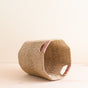 The LIKHA Abaca Octagon Basket, crafted from natural fibers, rests on its side with two pink-thread-wrapped handles. Its distinctive geometric form sits on a light wooden surface against a neutral background.