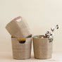 Three LIKHA Abaca Octagon Baskets with colored handles are displayed on a light surface against a beige background. One basket is tilted inside another, while the third holds cotton stems. All are ethically handcrafted from abaca fiber.