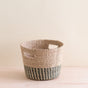 The LIKHA Abaca Tapered Basket is a Fair Trade handcrafted piece made from Manila hemp, with two rectangular handles and natural plus dark greenish-gray stripes at the bottom, shown on a light wooden surface against a neutral background.