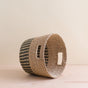 The LIKHA Abaca Tapered Basket, handwoven from sustainable natural fibers with two rectangular handles and a subtle striped pattern, rests on its side atop a light wooden surface against a beige background.