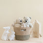 The LIKHA Abaca Tapered Basket, handwoven from Manila hemp, holds two teddy bears and a plush mouse. Nearby are two white ceramic bunny figurines and a decorative house, all arranged on a beige background.