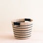 The LIKHA Abaca Tapered Basket, featuring black and natural stripes and two small black-wrapped handles, is crafted from durable Manila hemp fiber and sits on a light wooden surface against a plain background.