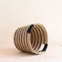 The LIKHA Abaca Tapered Basket, crafted from Manila hemp fiber, features bold black and natural stripes and is displayed on its side on a light wooden surface with a simple, light background.