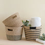 Two LIKHA Abaca Tapered Baskets, one with green stripes leaning inside the other. The black and natural striped basket holds rolled white towels and green foliage on a light surface.