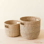 The LIKHA Abaca Round Bottom Basket, a nested set of 2, sits empty with handles on a light wooden surface against a cream background, showcasing sustainable storage in two sizes.