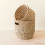 The LIKHA Abaca Round Bottom Basket, nested set of 2, made from natural fibers with rectangular cut-out handles, sits stacked on a light wooden surface against an off-white background—offering a stylish, sustainable storage solution.