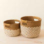 The Abaca Round Bottom Basket, a nested set of 2 by LIKHA, features handwoven Manila hemp with handles and a two-tone design—solid brown on top and a cream-brown braided pattern below—shown on a light wooden surface against a neutral backdrop.