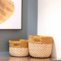 The LIKHA Abaca Round Bottom Basket, a nested set of 2 with handles and a two-tone natural and tan design, sits on a wooden surface in the corner next to framed abstract wall art.