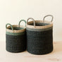 The LIKHA Abaca Cylinder Baskets, a nested set of 2 with handles, feature a color block pattern in dark green, beige, and light green. Handwoven from sustainable abaca, they rest on a light wooden surface against a neutral background.