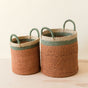 The LIKHA Abaca Cylinder Basket with handles, a nested set of 2, features ethically crafted Manila hemp fiber in brown with green and cream bands; both baskets sit side by side, one slightly larger than the other.